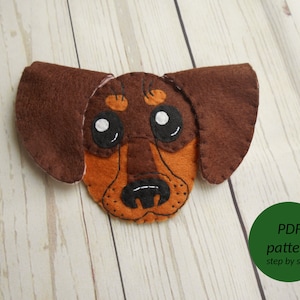 Puede incluir: Una cabeza de perro dachshund de fieltro con orejas marrones, pelaje naranja y marrón y ojos negros. El texto "PDF pattern step by step" está en un círculo verde.