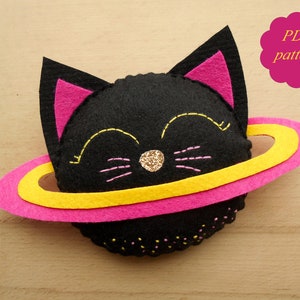 May include: A black felt cat with pink ears and a yellow and pink felt ring around it. The cat has a sparkly gold nose and pink embroidered whiskers. The image includes the text "PDF pattern".