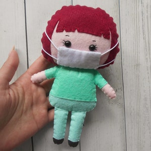 May include: A felt doll wearing a green shirt and pants with a white face mask. The doll has red hair and black eyes.