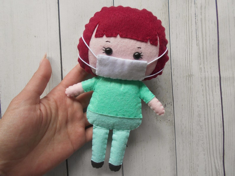 May include: A felt doll wearing a green shirt and pants with a white face mask. The doll has red hair and black eyes.