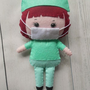 May include: A felt doll dressed as a doctor, wearing a green scrub top, pants, and a surgical cap. The doll has red hair, a white face mask, and black eyes.