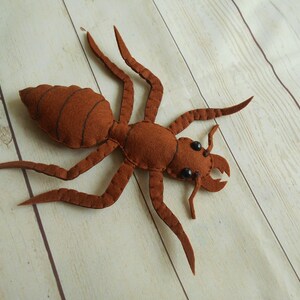 May include: A brown felt ant with black eyes. The ant is lying on its back with its legs spread out.