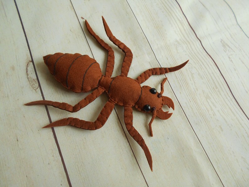 May include: A brown felt ant with black eyes. The ant is lying on its back with its legs spread out.