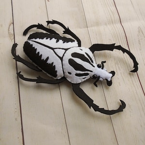 May include: A handmade felt beetle in black and white. The beetle has a white body with black markings and black legs and antennae. The felt insect is lying on a light wood surface.