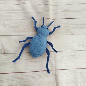 Puede incluir: Un juguete de insecto de fieltro azul hecho a mano con patas y antenas azul oscuro. El cuerpo es azul claro con detalles cosidos. El juguete está sobre una superficie de madera clara.