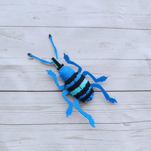 May include: A blue and black felt bug with long legs and a striped body. The bug is lying on a white wooden surface.