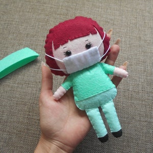 May include: A felt doll wearing a green scrub top and pants, a white face mask, and red hair. The doll is holding a small green felt rectangle.