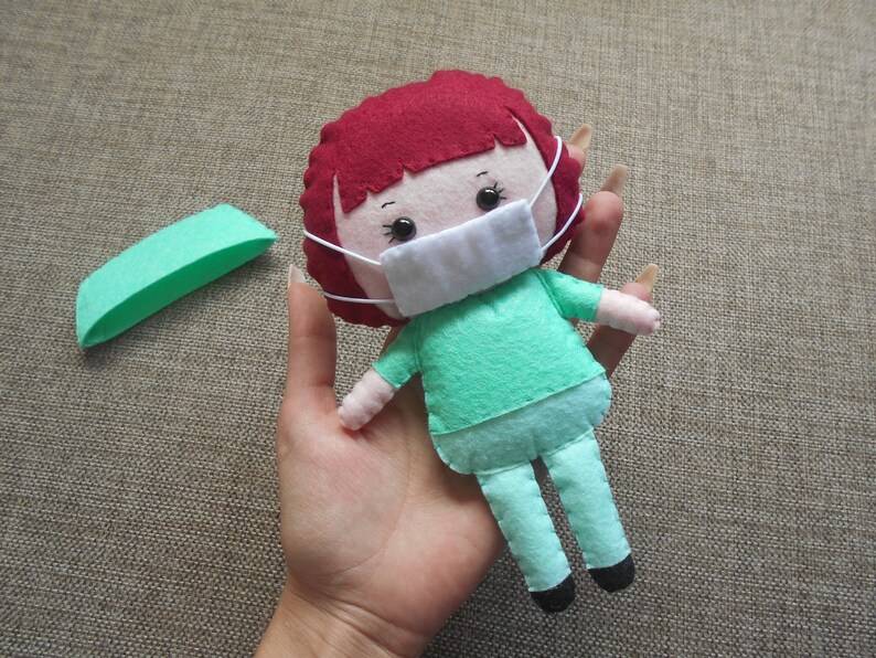 May include: A felt doll wearing a green scrub top and pants, a white face mask, and red hair. The doll is holding a small green felt rectangle.
