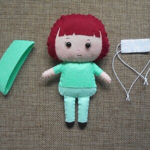 May include: A felt doll with red hair, wearing a green shirt and pants, and a white face mask. The doll is holding a green felt rectangle.