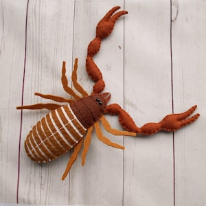 Puede incluir: Un juguete de fieltro hecho a mano con forma de escorpión en tonos marrones y naranjas. El escorpión tiene un cuerpo segmentado con rayas blancas, grandes pinzas y múltiples patas. El juguete está diseñado para parecerse a un escorpión real.