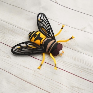 Puede incluir: Una abeja de fieltro con rayas negras y amarillas y alas transparentes. La abeja está tumbada sobre una superficie de madera blanca.