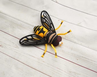 May include: A felt bee with black and yellow stripes and clear wings. The bee is lying on a white wooden surface.