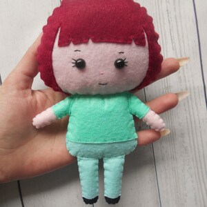 May include: A felt doll with red hair, a pink face, black eyes, a green shirt, and blue pants.