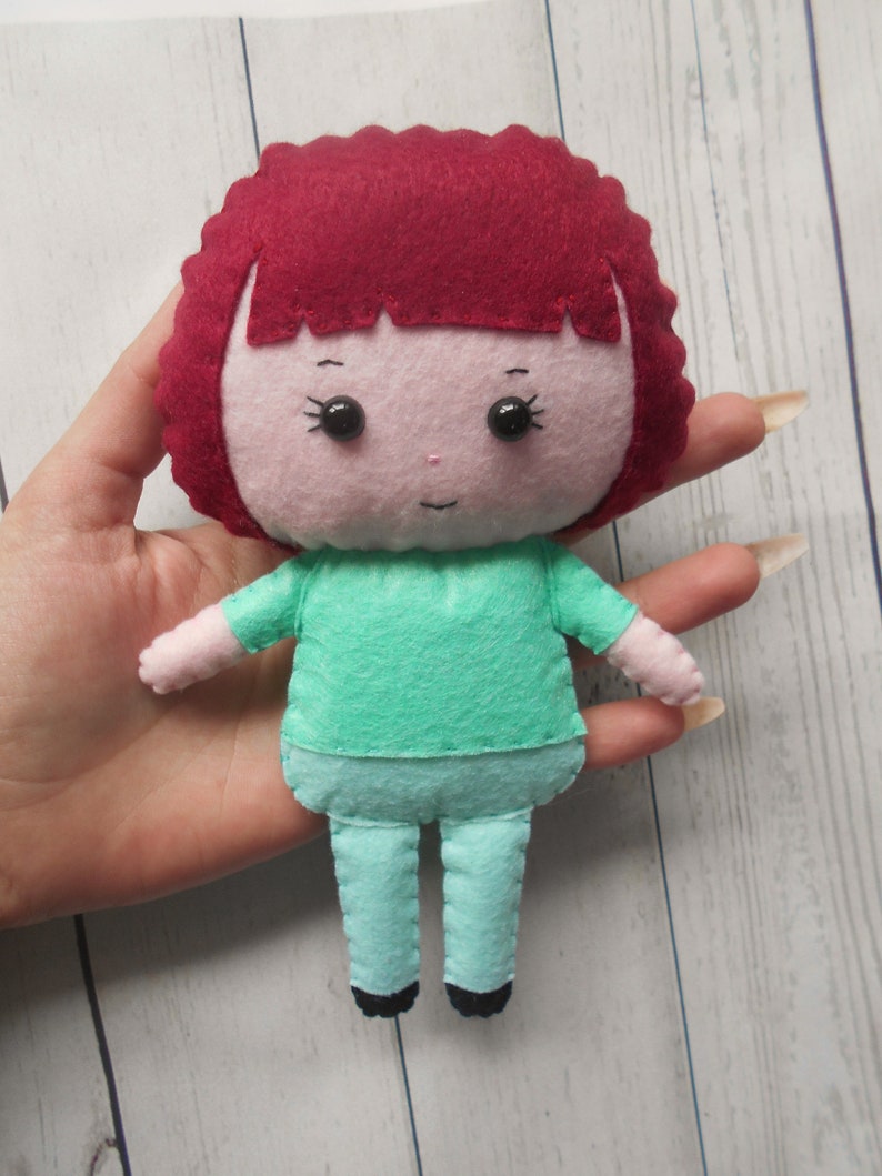 May include: A felt doll with red hair, a pink face, black eyes, a green shirt, and blue pants.