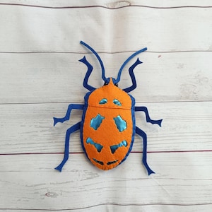 Puede incluir: Un insecto de fieltro hecho a mano en naranja y azul. El insecto tiene patas, antenas y detalles azules en su cuerpo naranja. El insecto de fieltro está sobre una superficie clara con vetas de madera.