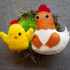 K&ouml;nnte beinhalten: Zwei Filz-Spielzeug-H&uuml;hner in einem Nest. Das gr&ouml;&szlig;ere Huhn ist wei&szlig; mit braunen Fl&uuml;geln und einem roten Kamm. Das kleinere Huhn ist gelb mit einem roten Kamm.