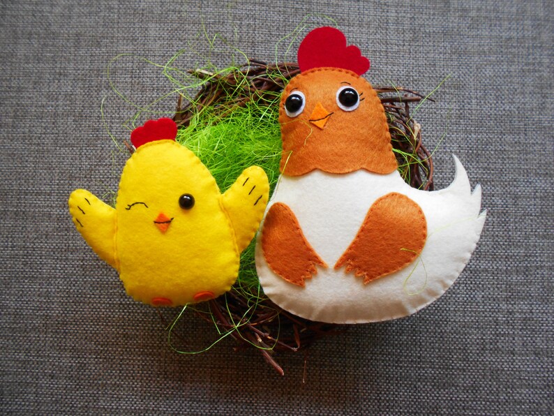 K&ouml;nnte beinhalten: Zwei Filz-Spielzeug-H&uuml;hner in einem Nest. Das gr&ouml;&szlig;ere Huhn ist wei&szlig; mit braunen Fl&uuml;geln und einem roten Kamm. Das kleinere Huhn ist gelb mit einem roten Kamm.