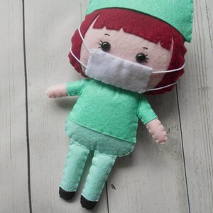May include: A felt doll dressed as a doctor, wearing a green scrub top, green pants, a green hat, and a white face mask. The doll has red hair and black eyes.