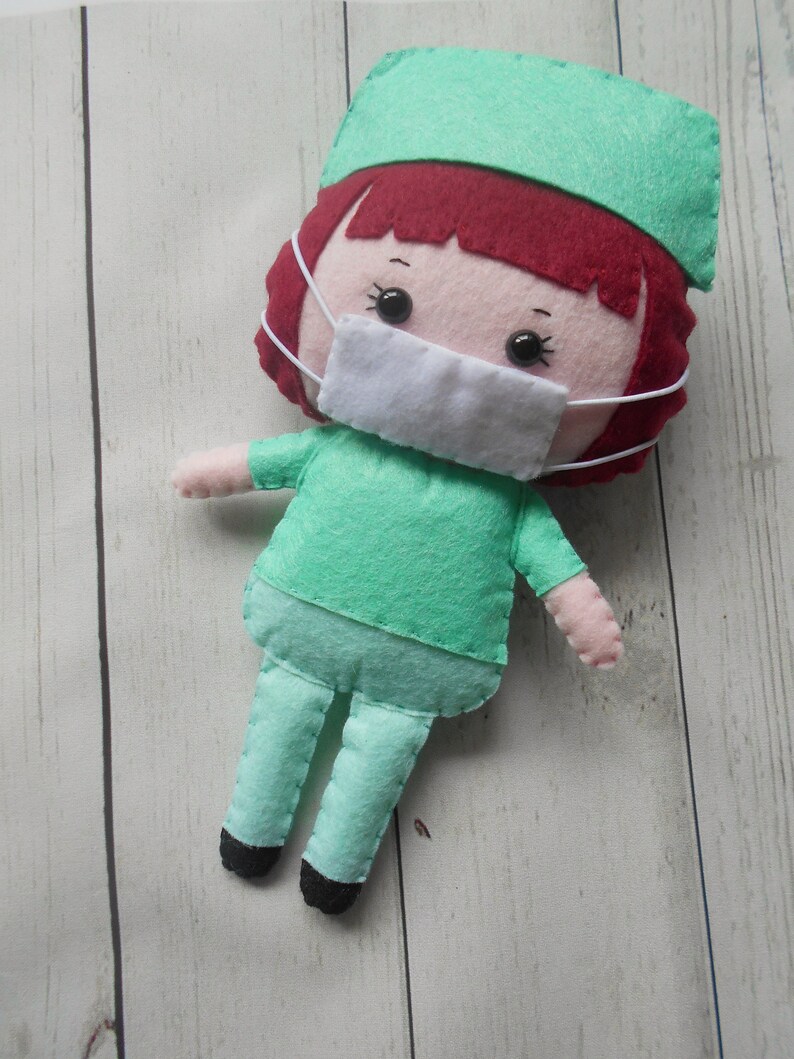 May include: A felt doll dressed as a doctor, wearing a green scrub top, green pants, a green hat, and a white face mask. The doll has red hair and black eyes.