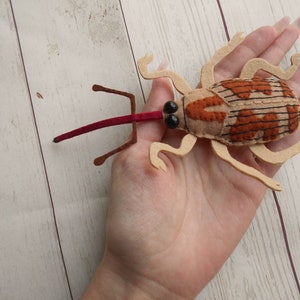 May include: A handmade felt insect toy with a tan body, brown and orange markings, and a long red proboscis. The toy has black bead eyes and tan legs and antennae. The insect is shown on a light wood grain surface.
