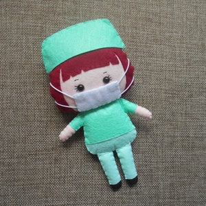 May include: A felt doll wearing a green scrub cap and a white face mask. The doll has red hair and a green scrub top and pants.