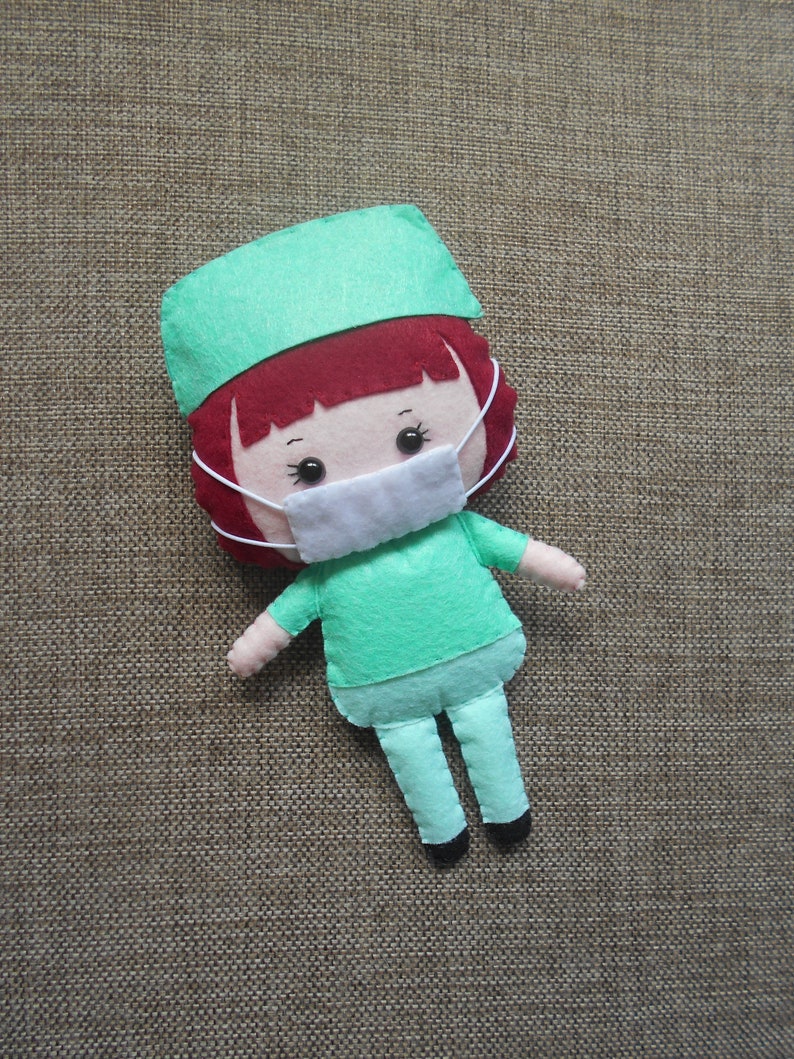 May include: A felt doll wearing a green scrub cap and a white face mask. The doll has red hair and a green scrub top and pants.
