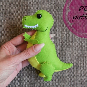 May include: Green felt dinosaur toy with black eyes and a yellow belly. The toy is being held by a hand. The text "PDF pattern" is visible in the background.