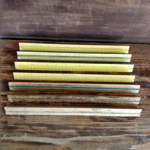May include: A collection of seven triangular architectural scales and rulers arranged horizontally. The set includes four yellow plastic scales with black markings, one wooden scale with a green stripe, and two weathered wooden rulers with visible markings.