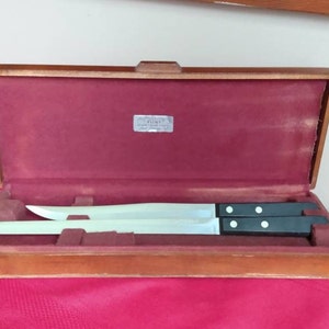May include: A vintage carving set in a wooden case. The set includes two long, stainless steel knives with black handles. The interior of the case is lined with a burgundy velvet material. A small silver label is visible inside the case.