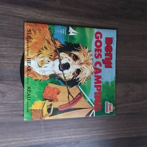 May include: A vintage record featuring a cartoon illustration of a dog named Benji going camping. The record cover is blue and white with the text "Benji Goes Camping" in red and white. The dog is holding a stick and there is a tent and a hot dog in the background.