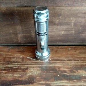 May include: A vintage chrome flashlight with a ribbed body and a rectangular label that reads "RANGER U.S.A." The flashlight has a switch and a circular top and base. The flashlight is on a wooden surface.