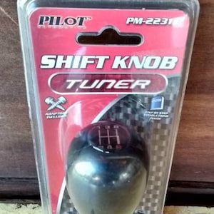 May include: A black shift knob tuner with a red, white, and black packaging. The packaging has the text "PILOT" and "SHIFT KNOB TUNER" on it. The shift knob has a 5-speed pattern.