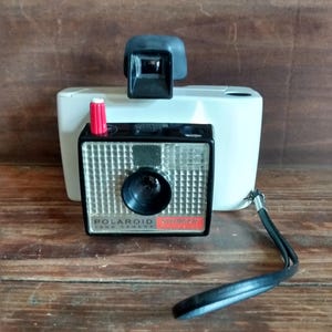 May include: Vintage Polaroid Swinger Land Camera with a black lens, silver grid, and red accents. The camera has a white body and a black viewfinder. A black wrist strap is attached. The camera is on a wooden surface.