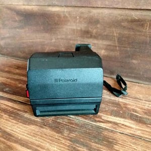 May include: A black Polaroid instant camera with the brand name printed on the front. The camera has a red button on the side and a black strap. The camera is sitting on a wooden surface.