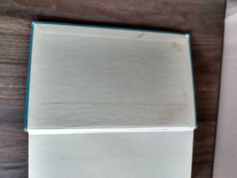 K&ouml;nnte beinhalten: Ein tealblaues Hardcover-Buch mit wei&szlig;em R&uuml;cken und wei&szlig;en Seiten. Das Buch ist geschlossen und liegt flach auf einer Holzoberfl&auml;che.