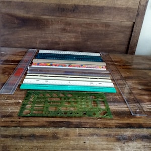 May include: A collection of various rulers and a drafting template. The set includes several straight rulers in clear, white, tan, and green plastic, along with one featuring a Snoopy graphic. A green plastic drafting template with geometric cutouts.