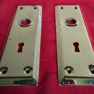 May include: Two antique brass door plates with keyholes and rounded edges. The plates have a worn, aged finish.