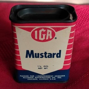 May include: A red and white metal container with a black lid. The container is labeled "IGA Mustard" and has a net weight of 1 1/4 ounces. It is packed for Independent Grocers Alliance Distributing Co., Inc. in Chicago, Illinois, USA.
