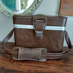 May include: A brown leather carrying case with a silver trim and a shoulder strap. The case has a metal latch and a flap with a metal buckle.