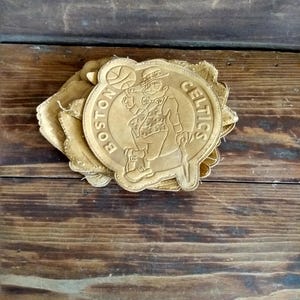 May include: A stack of tan leather patches featuring the Boston Celtics logo. The logo depicts a leprechaun holding a basketball, surrounded by the words "BOSTON CELTICS" in a circular design. The patches are on a wooden surface.
