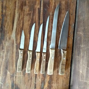 May include: A set of six kitchen knives with wooden handles and stainless steel blades. The knives vary in size, from a small paring knife to a large carving knife. The handles are a light brown color, and the blades are silver.