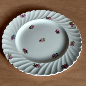 May include: White ceramic plate with a scalloped edge and a floral design. The plate has a swirl pattern and features small pink, yellow, and blue flowers.