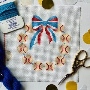 May include: A white canvas with a red, white, and blue bow at the top and a wreath of baseballs around the bow. The canvas is surrounded by gold and white confetti.