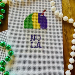 Puede incluir: Un lienzo blanco con una máscara de Mardi Gras morada, verde y amarilla con una pluma roja y blanca. Las palabras "NOLA" están bordadas en morado debajo de la máscara.