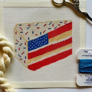 May include: A needlepoint canvas with a design of a red, white, and blue American flag on a yellow cake. The canvas is surrounded by a white border. A skein of white embroidery floss and a skein of blue embroidery floss are visible in the image. The embroidery floss is labeled "Tille essen Silk 50: Mer 10 yards LO YING 80 500500108 info@planetearth Dry Block".