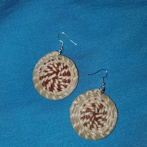 May include: A pair of handcrafted earrings with a woven design. The earrings feature a circular shape with a spiral pattern in shades of brown, beige, and green. The earrings have silver-colored hooks.
