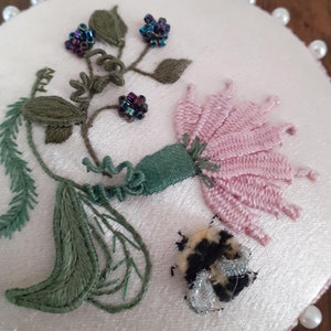 Pode incluir: Uma flor rosa com folhas verdes e contas de cor púrpura iridescente é bordada em um círculo de tecido branco com uma borda de pérolas. Um pequeno zangão preto e amarelo é bordado abaixo da flor.