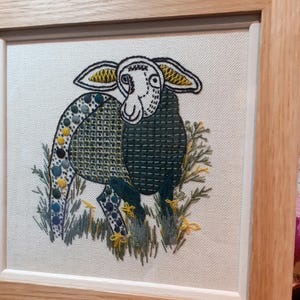 May include: Embroidered artwork of a sheep in a wooden frame. The sheep has a white face, large ears with yellow accents, and a dark green body with blue and yellow details. The artwork is set against a white background with embroidered grass.