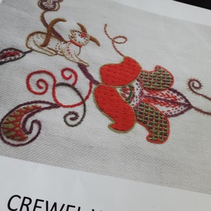 May include: Close-up of a crewel work embroidery design on a light fabric. The design features a whimsical goat-like creature, red flowers, and swirling vines in shades of red, green, brown, and purple. The text "CREWEL WORK" is visible.