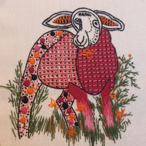 May include: Embroidered artwork of a sheep with a white face and ears with orange accents. The sheep's body is red with orange and black polka dots and a cross-stitch pattern. Green grass and orange flowers surround the sheep.